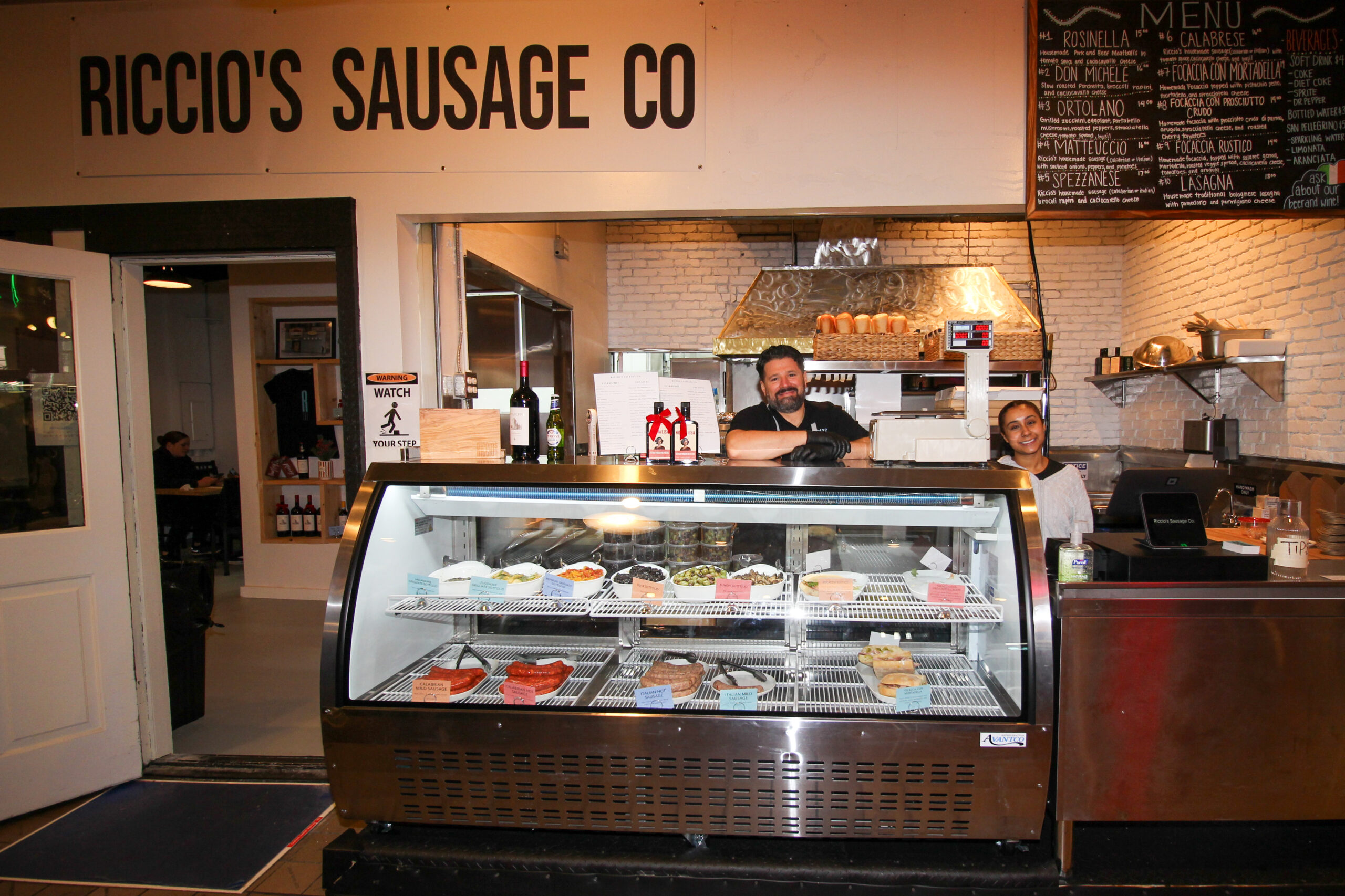 man behind deli counter showcasing sandwiches and food items